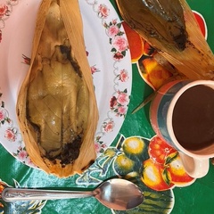Mole Tamal made by Doña Inés (© Daniela Sclavo). An opened tamale sits on a floral-patterned plate next to a cup of coffee. These and several pieces of cutlery are photographed from above, with a colorful tablecloth underneath.