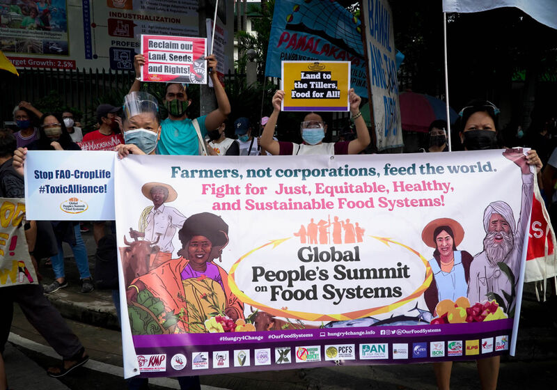 A crowd of protesters carry banners reading "Farmers, not corporations, feed the world. Fight for just, equitable, healthy and sustainable food systems!"