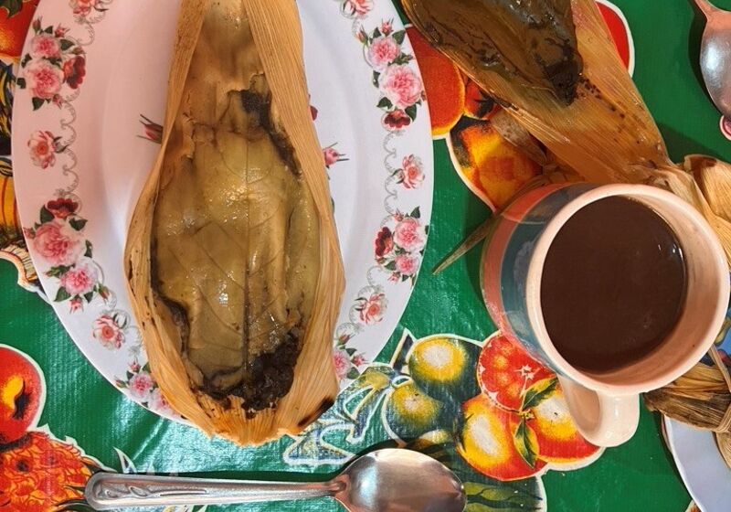 Mole Tamal made by Doña Inés (© Daniela Sclavo). An opened tamale sits on a floral-patterned plate next to a cup of coffee. These and several pieces of cutlery are photographed from above, with a colorful tablecloth underneath.