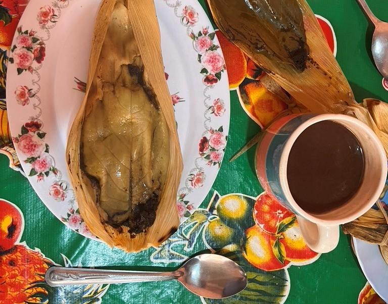 Mole Tamal Mole Tamal made by Doña Inés (© Daniela Sclavo). An opened tamale sits on a floral-patterned plate next to a cup of coffee. These and several pieces of cutlery are photographed from above, with a colorful tablecloth underneath.