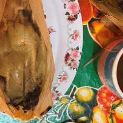 Mole Tamal made by Doña Inés (© Daniela Sclavo). An opened tamale sits on a floral-patterned plate next to a cup of coffee. These and several pieces of cutlery are photographed from above, with a colorful tablecloth underneath.